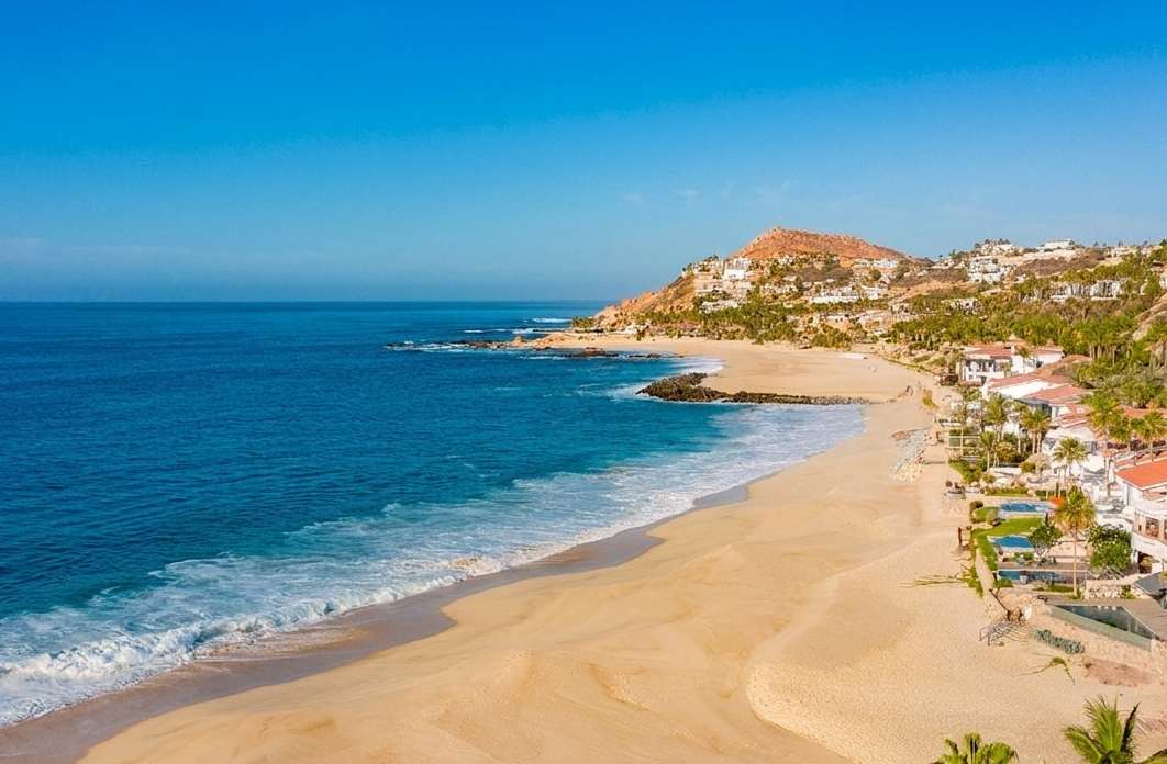 A vibrant, high-angle aerial view of a sweeping crescent beach with golden sand meeting a deep blue ocean. Bright white surf rolls onto the shoreline, while the right side of the frame features upscale coastal villas with red-tiled roofs, swimming pools, and manicured lawns lined with palm trees. In the distance, a rugged, sun-drenched hill dotted with luxury homes overlooks a rocky point jutting into the sea under a clear, brilliant blue sky.